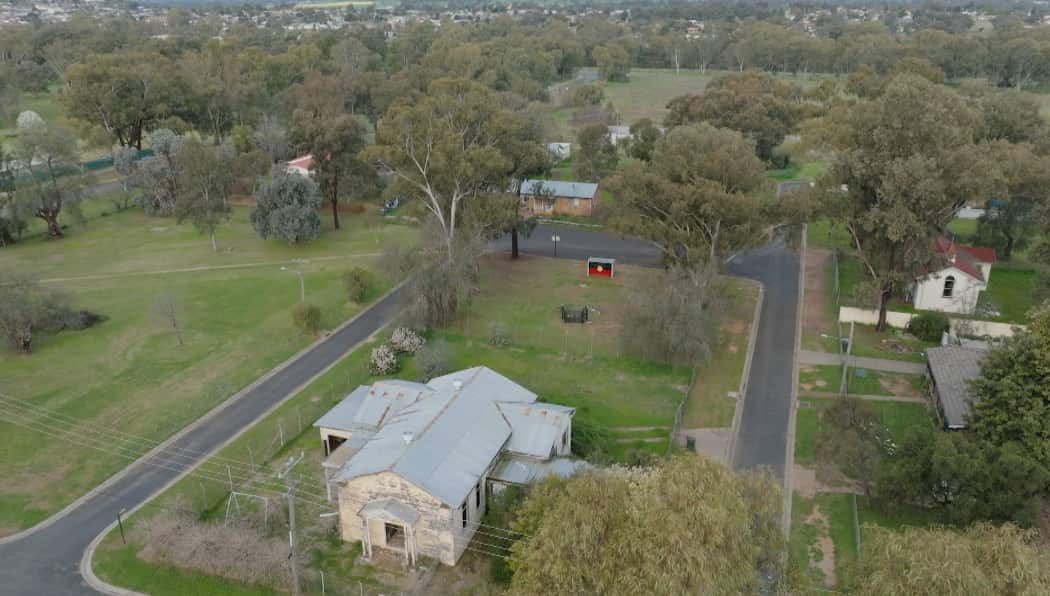 Aerial shot of Erambie Mission, also known as 32 acres for its size, including green parklands with a white wood building in the foreground and an Aboriginal flag painted on a bus stop in the background.