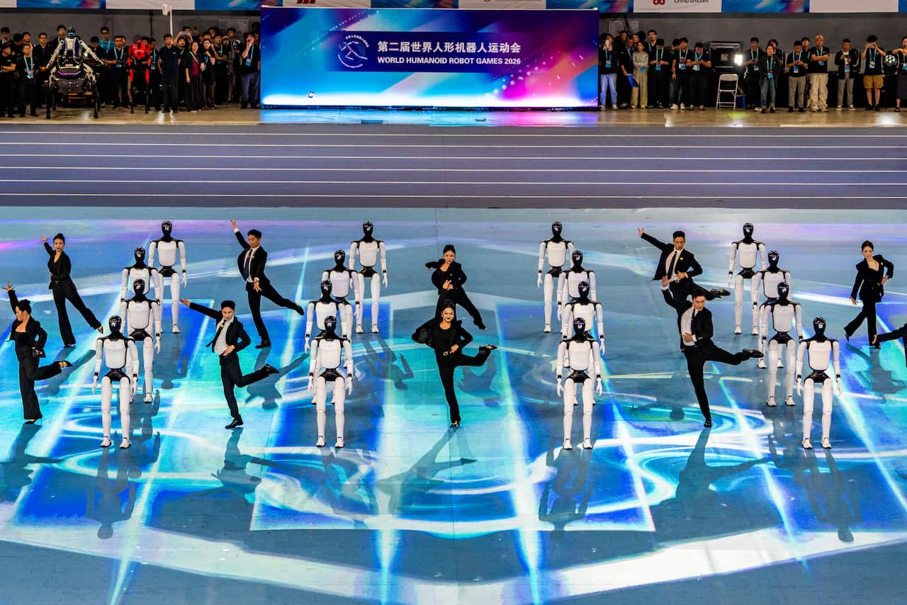 A group of dancers and human-like robots stand in rows in a stadium