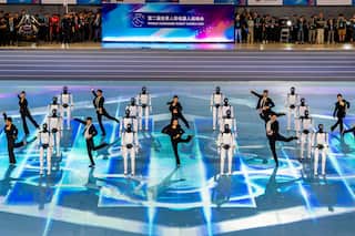 A group of dancers and human-like robots stand in rows in a stadium