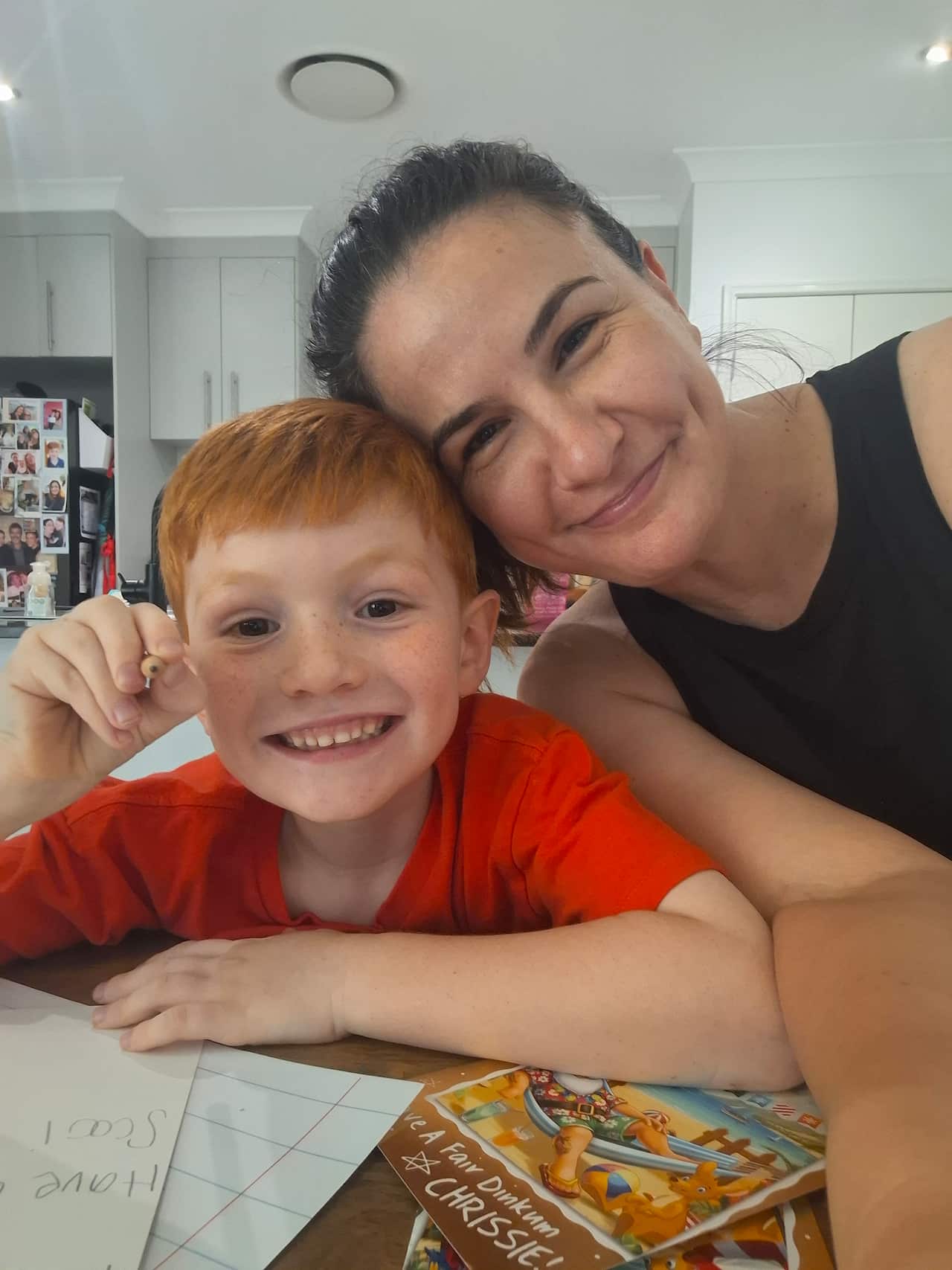 A mother and her young son smile together at the camera, with a kitchen in the background.