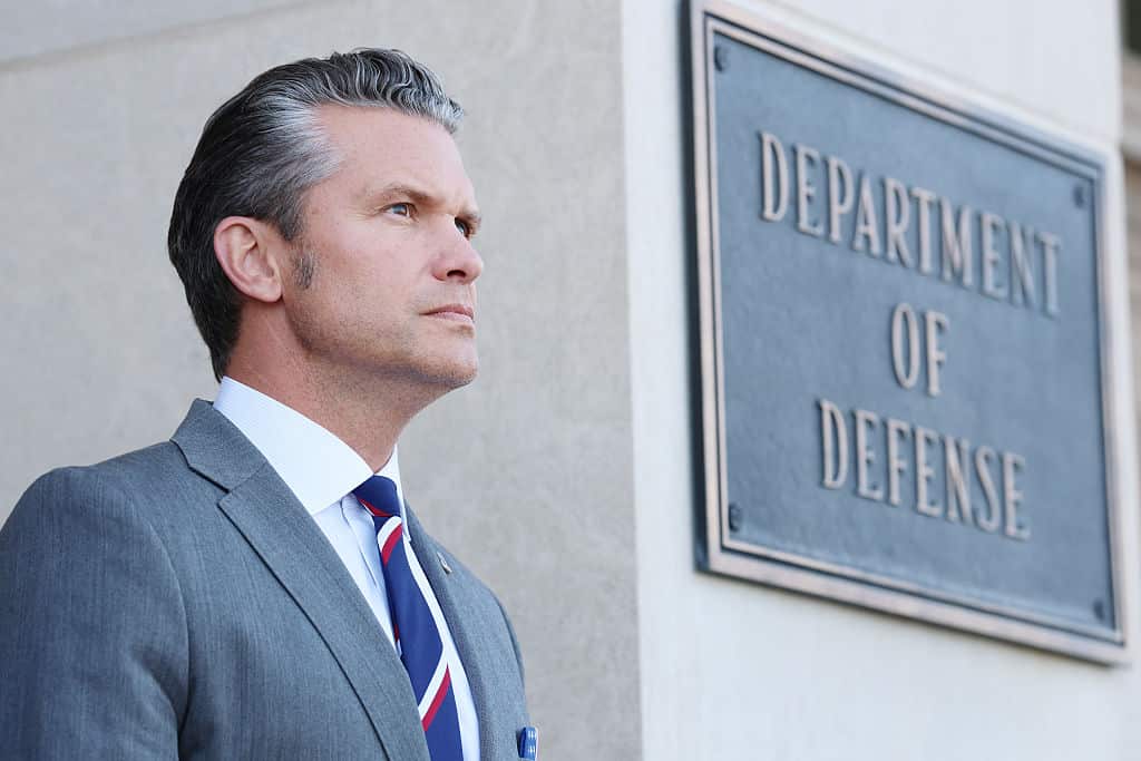 A man in a grey suit outside a building with a sign 'Department of Defense' over his shoulder.