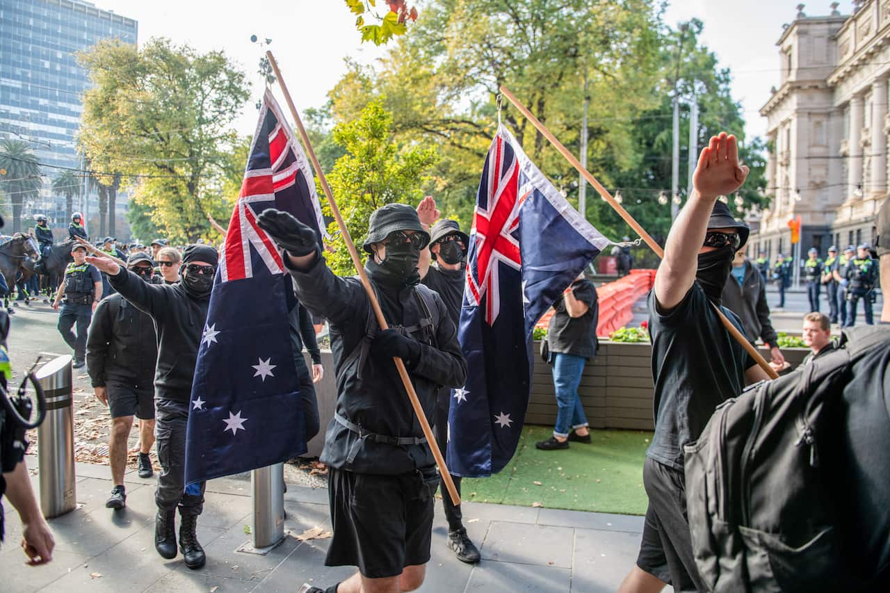 People dressed in black, with black masks covering their faces and holding Australian flags, salute as they walk. 