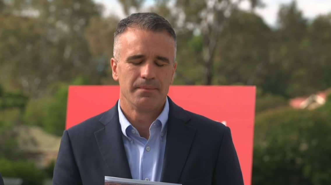 A medium shot of a man in a navy blazer and light blue shirt standing outdoors with his eyes closed, positioned in front of a red sign and a blurred background of trees.