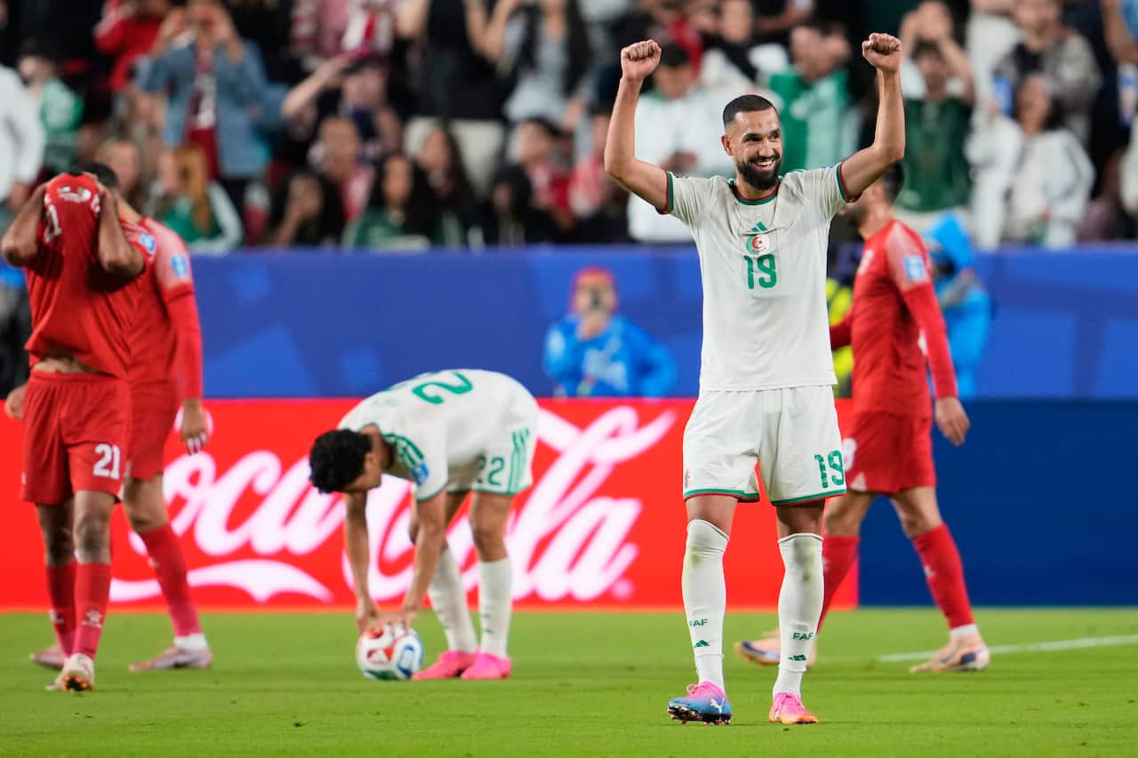 Five footballers on a green pitch, two dressed in white and two in red. One of the footballers dressed in white raises his hands and smiles in celebration.