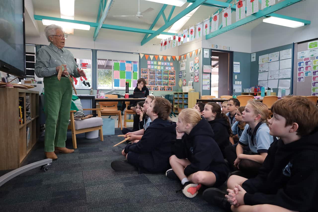 Wearing green pants and a top, an elderly woman shows a classroom of young primary school students a boomerang.