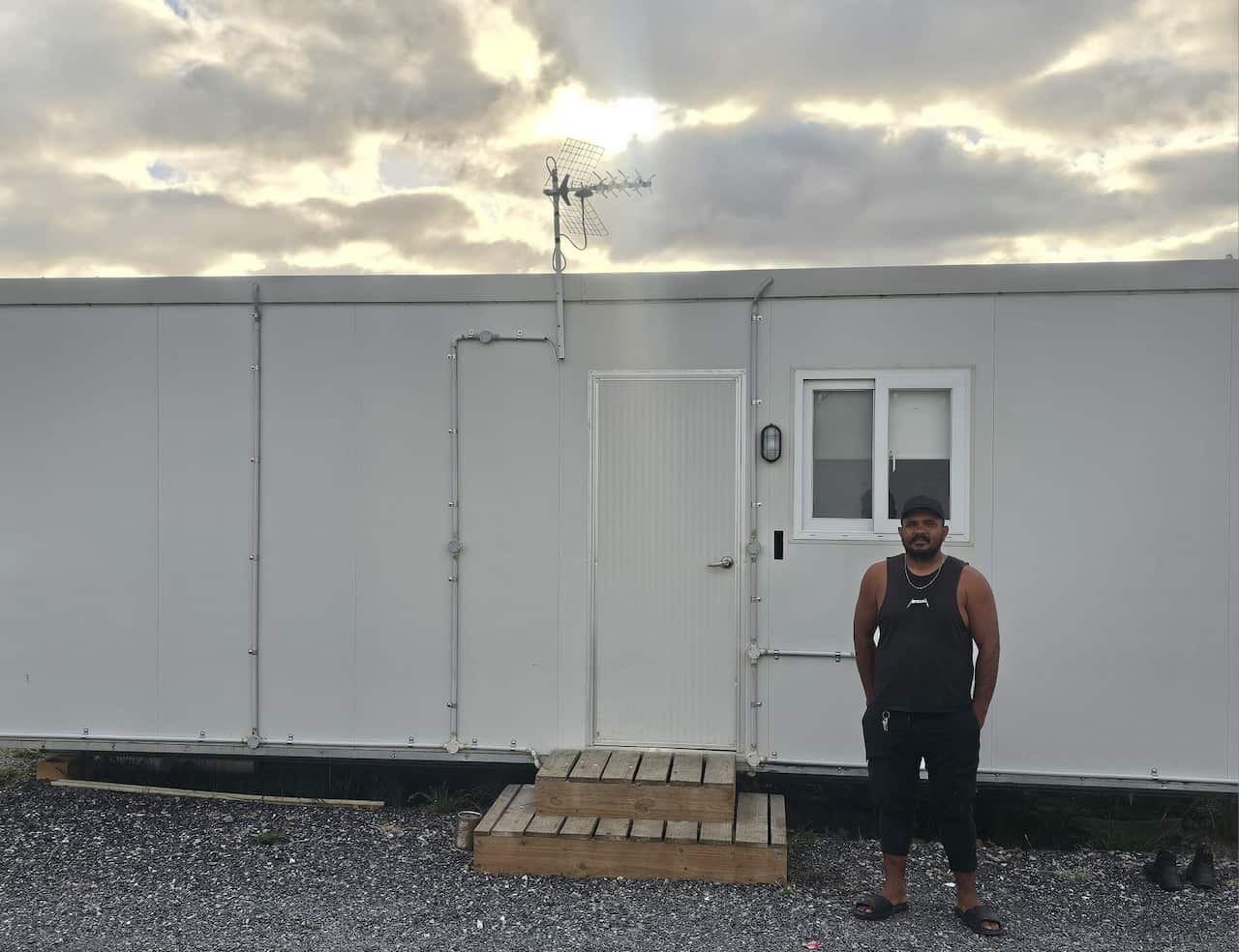 A man wearing a black singlet and trackpants stands outside a white caravan.