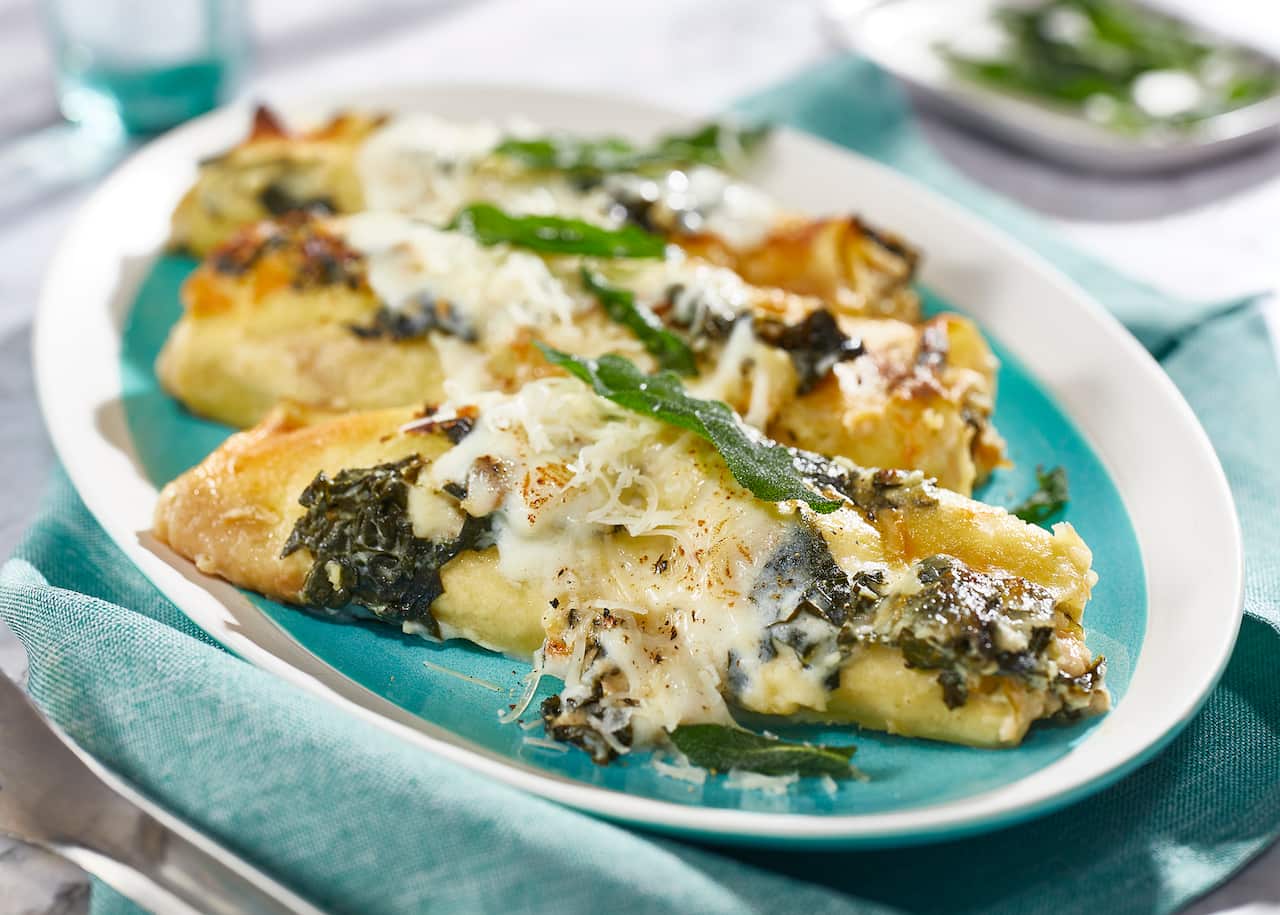Three cannelloni, topped with greens and cheese, sit on a blue platter with a white rim. The platte sits on a cloth napkin in the same shade of blue. 