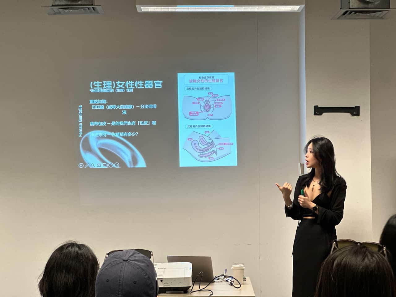 A woman in a black dress is giving a presentation to the audience members seated in a room.