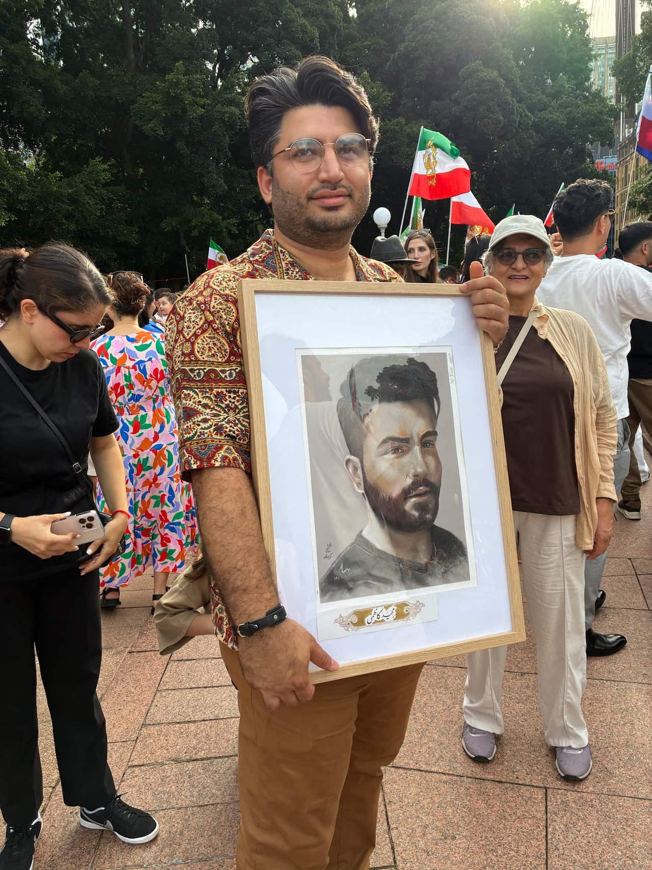 A man in a patterned shirt stands in a crowded outdoor area holding a large framed portrait of another man, with several people and Iranian flags visible in the background.