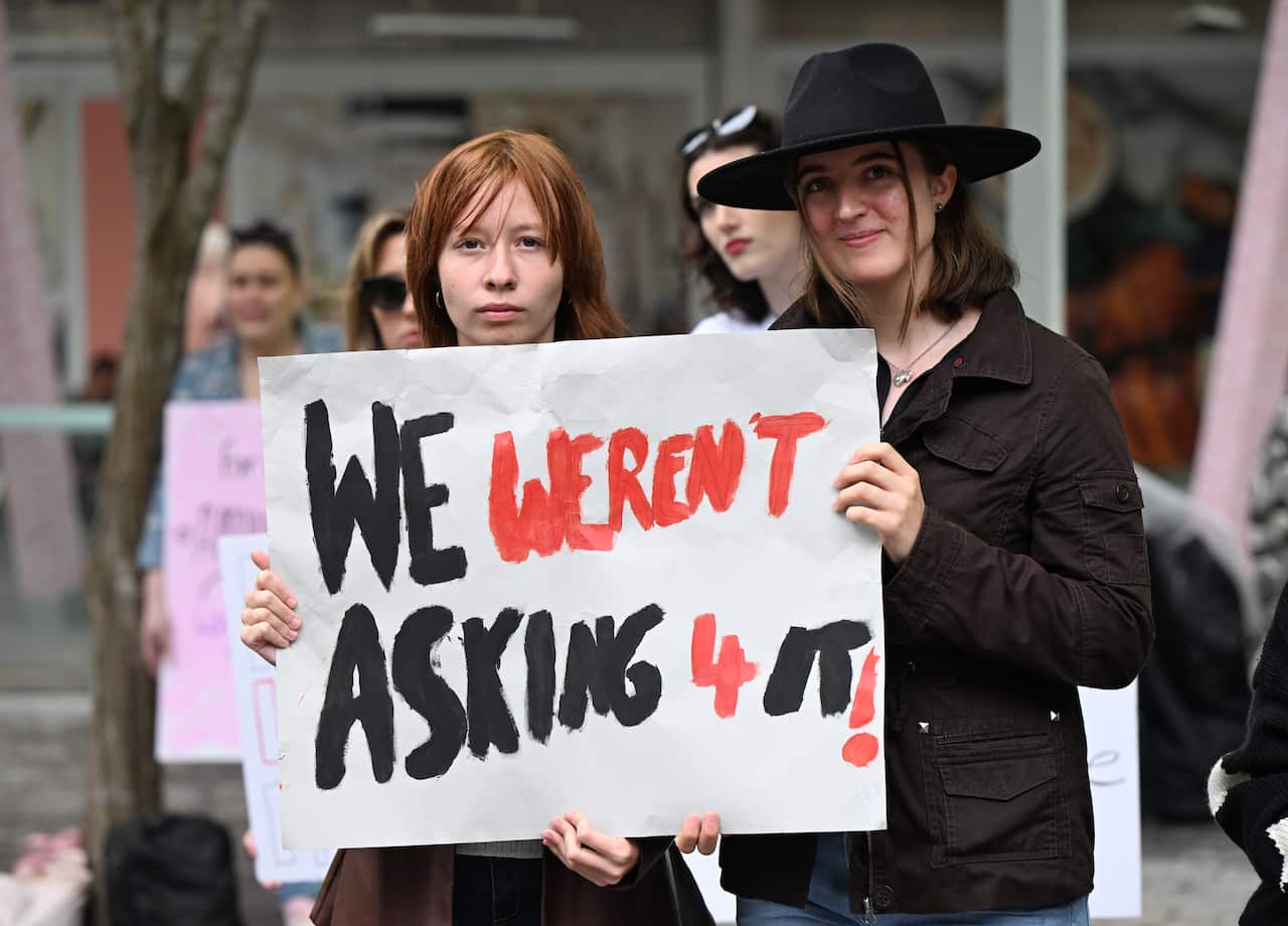Two women hold a sign that reads "WE WERENT ASKING 4 IT" at a protest.