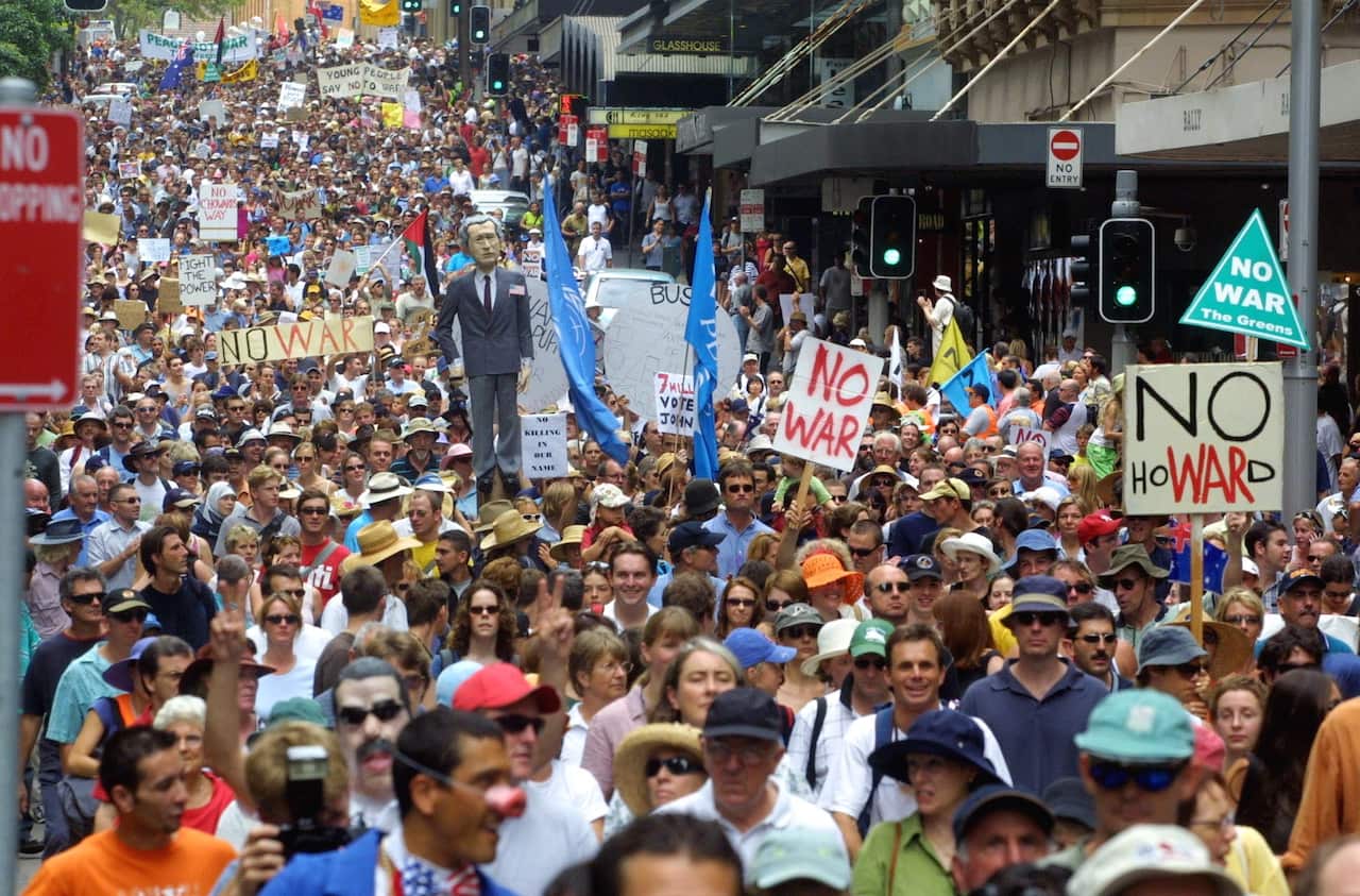 Demonstrators marching down a street. Some are holding placards that read "NO WAR". 