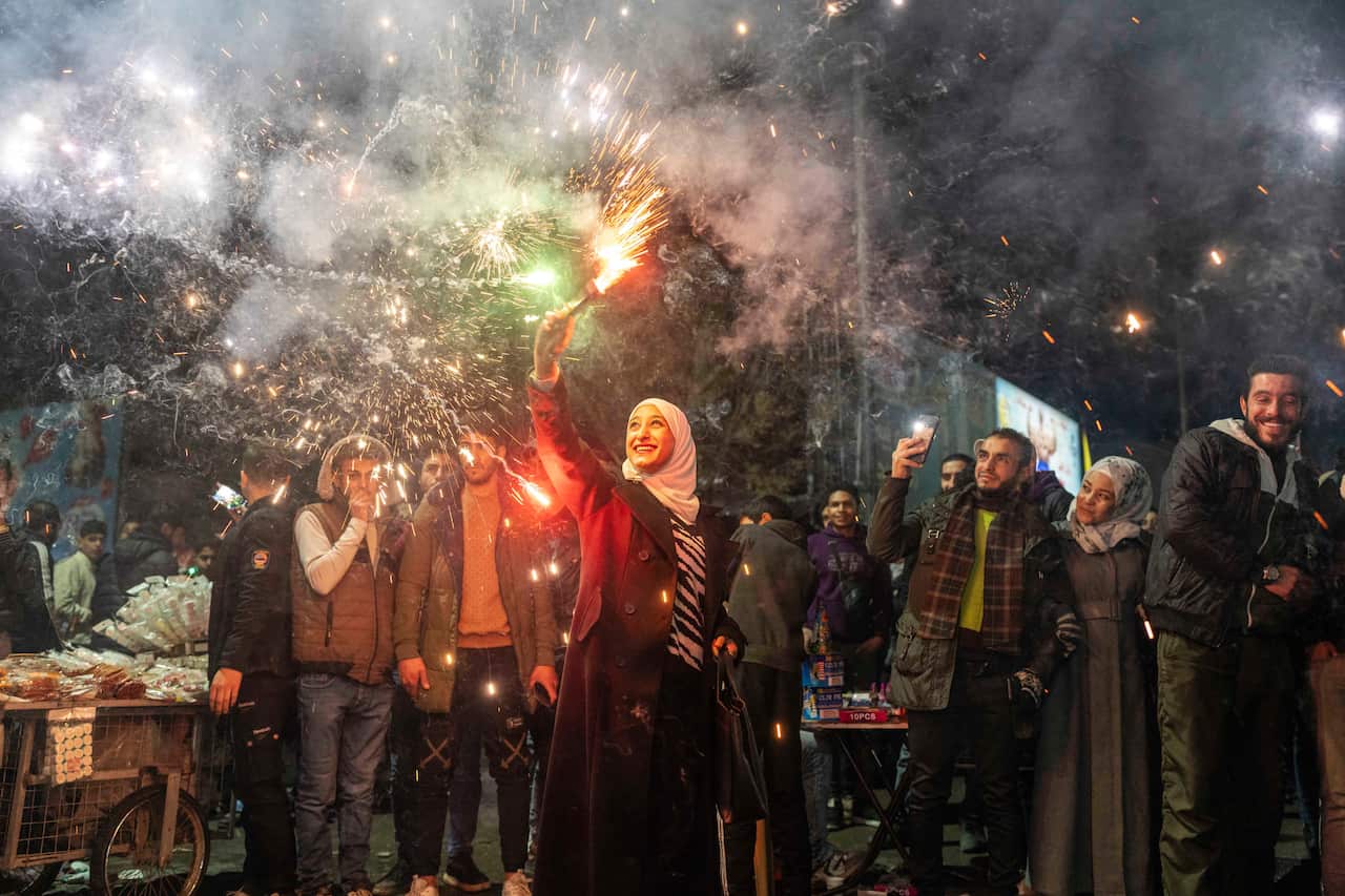 A woman holds a sparkler as she stands amidst a crowd, with smoke filling the sky around them.