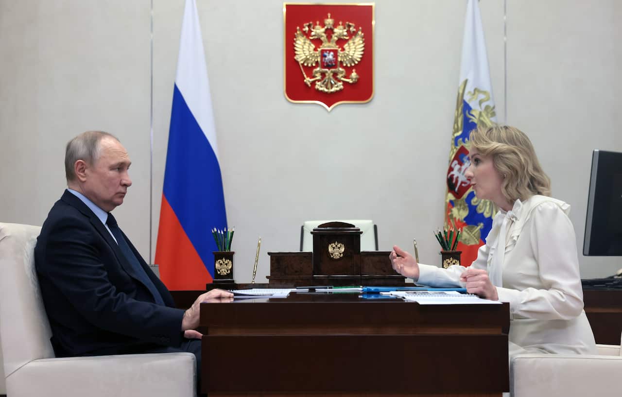 A man and a woman sit at a table with flags in the background.