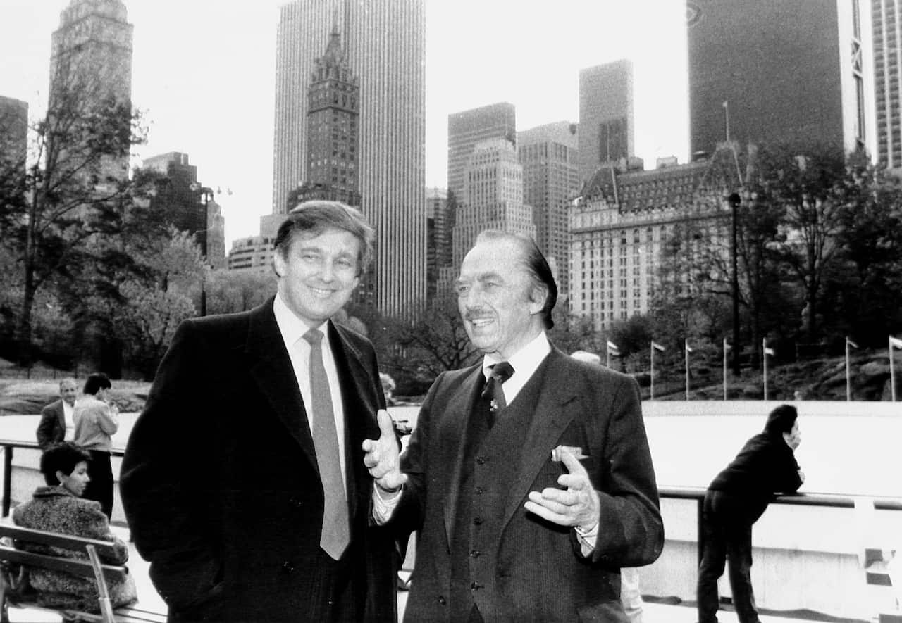 Donald Trump stands alongside his father, Fred Trump, in an open area with trees in the background. Several people can be seen behind them, and tall buildings are visible further away in the distance.
