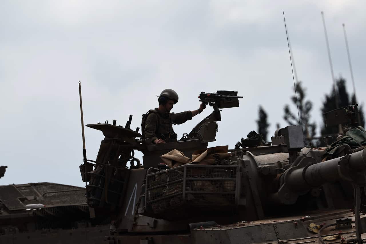 A soldier wearing a helmet and headset stands in the hatch of a military tank, gripping a mounted machine gun against an overcast sky.