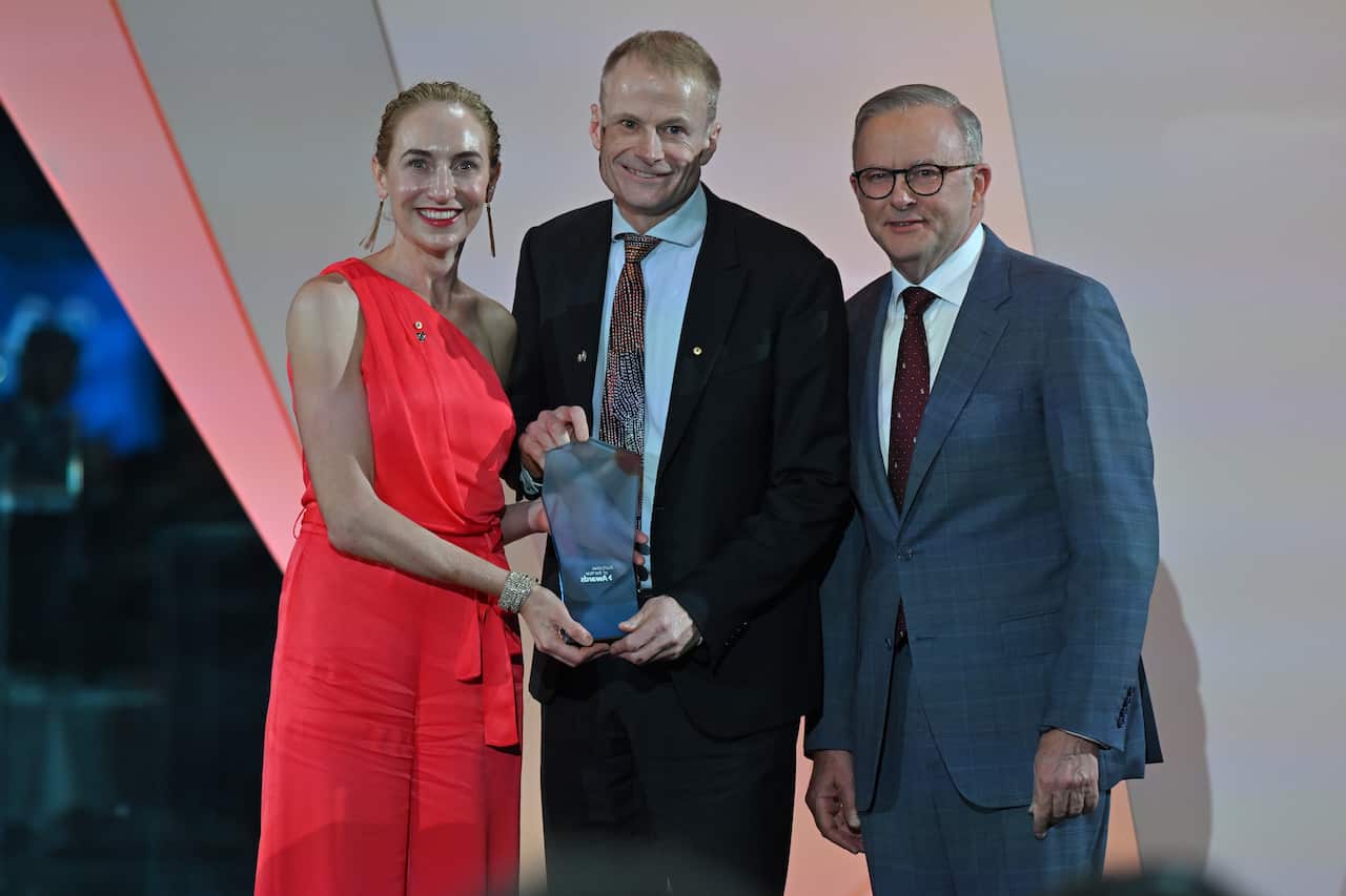 A woman in a red dress and a man in a suit hold an award while posing with Prime Minister Anthony Albanese.