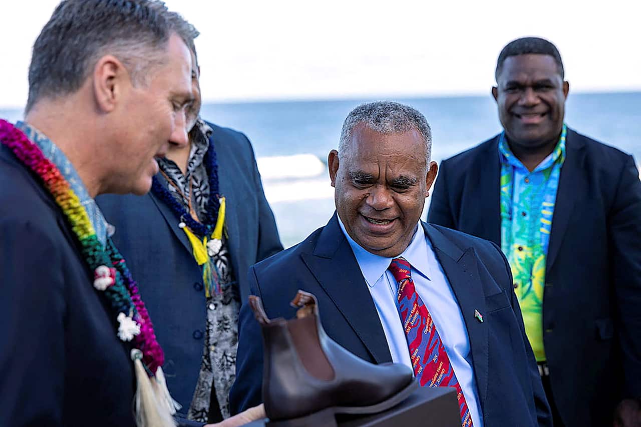 A man in a suit presents a shoe to another man in a suit, with two men standing behind them.
