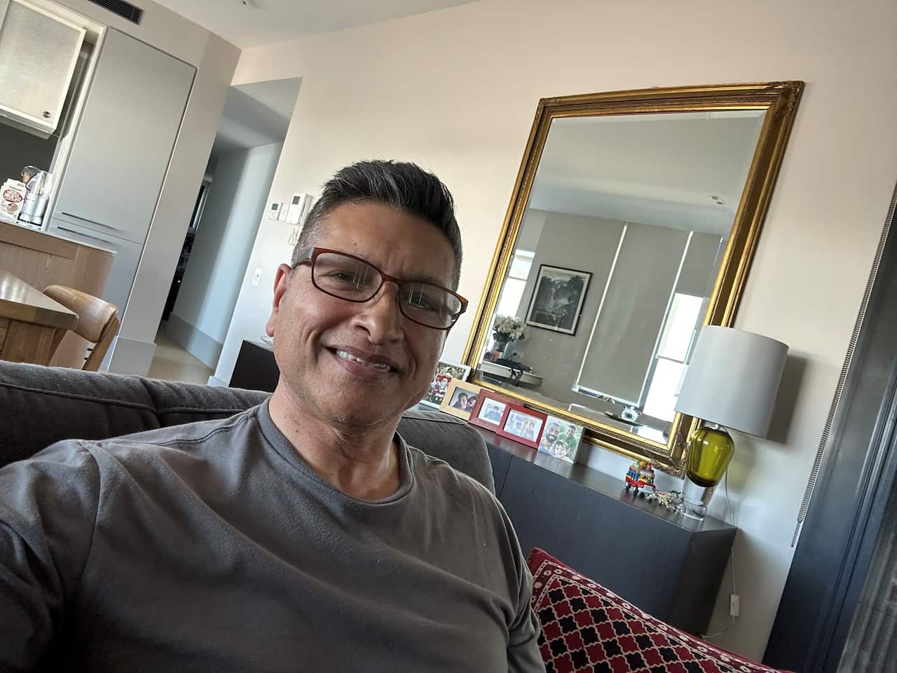 Alan sits on a lounge in a grey t-shirt and glasses, smiling into the camera