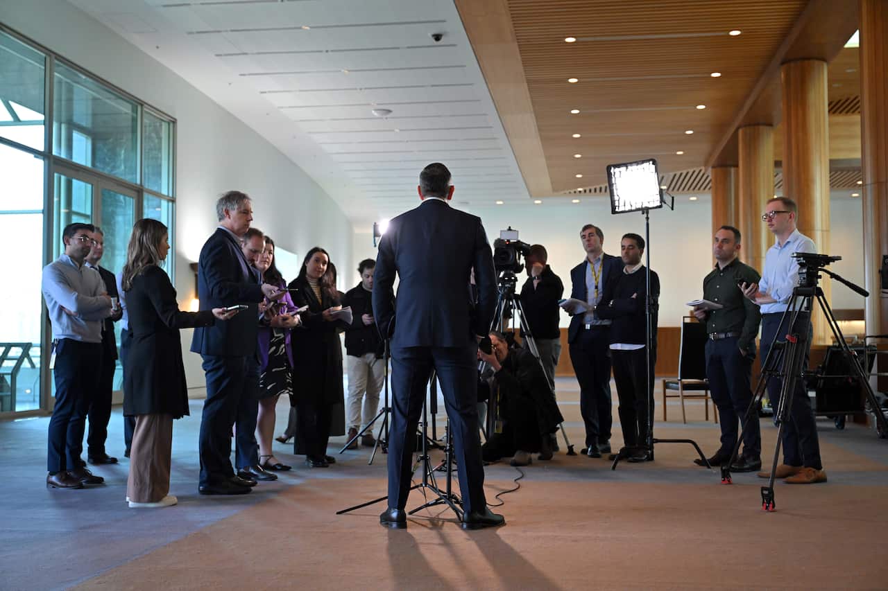 A man in a blue suit with his back turned in front of a row of people and cameras.