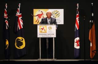 An older white man standing before a podium which has signs on it saying 'Bidding Nation Australia' and cartoon kangaroos and the Football Federation Australia logo and text