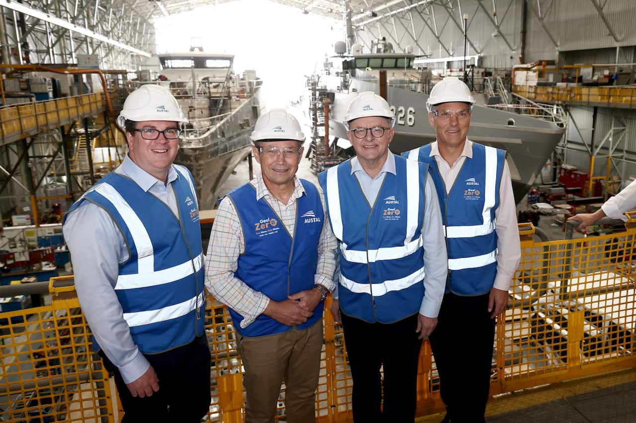 Four men wearing hard hats and blue and white vests stand in a shipyard. They are posing for a photo in front of a partially constructed naval vessel with the number 226 visible on its hull.
