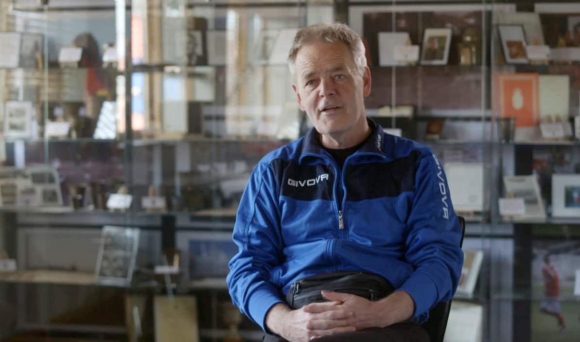 A man is interviewed in a room full of sporting memorabilia in the background.jpg