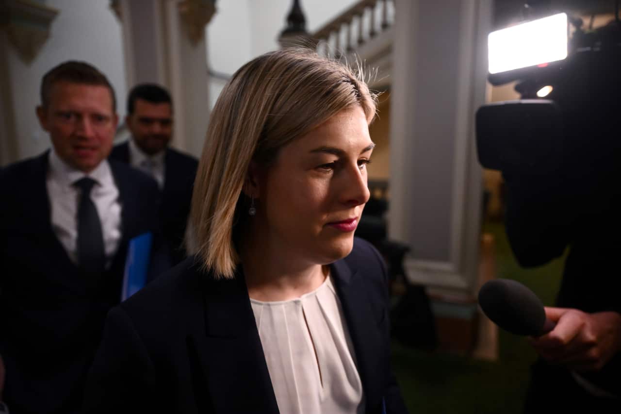 A woman with short blonde hair and wearing an off-white blouse and navy jacket inside.