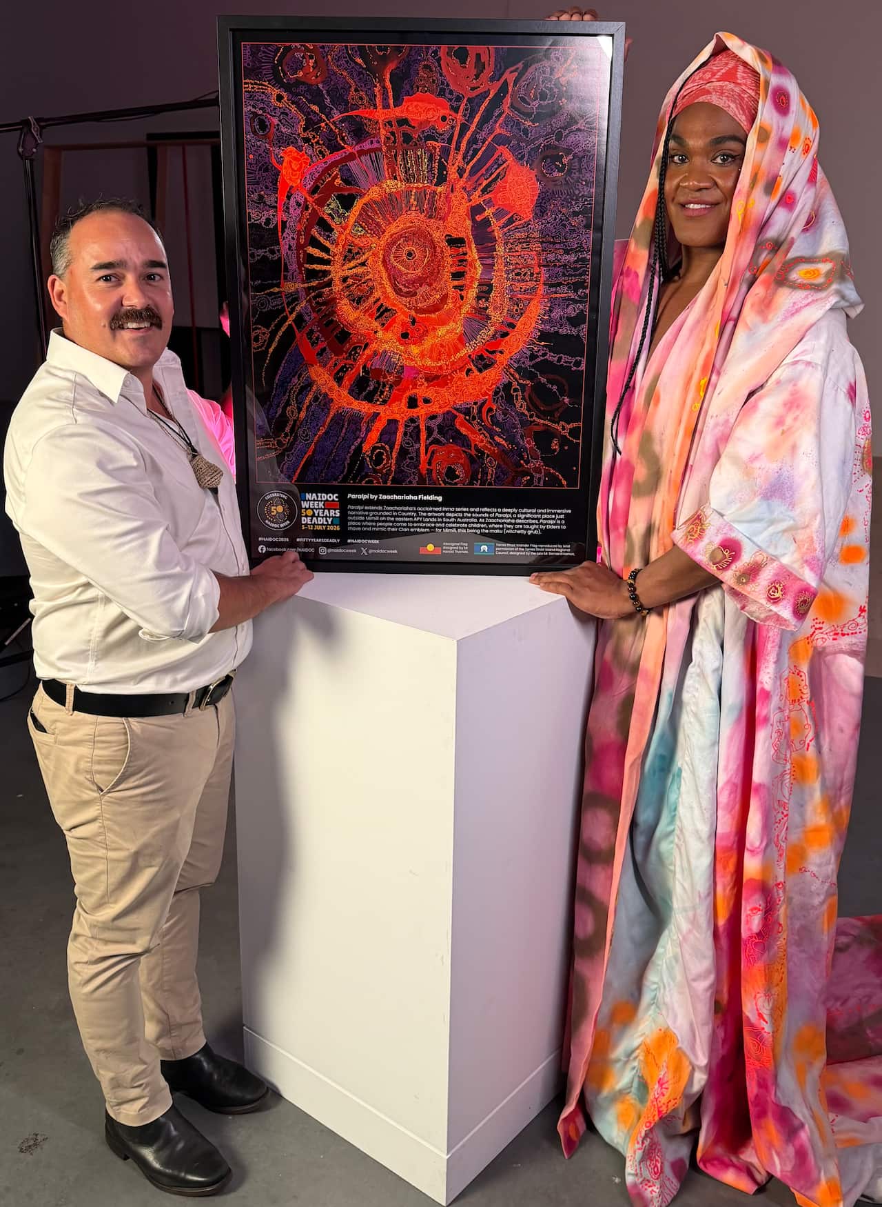 An artist wearing pastel clothing and a man wearing formal clothing hold up a framed artwork.