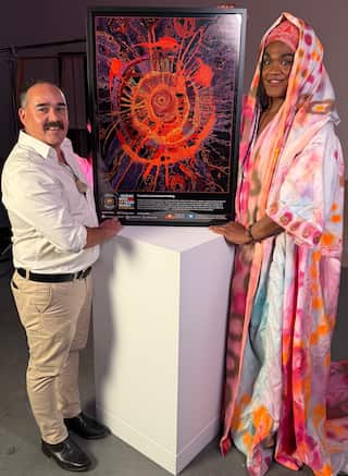 An artist wearing pastel clothing and a man wearing formal clothing hold up a framed artwork.