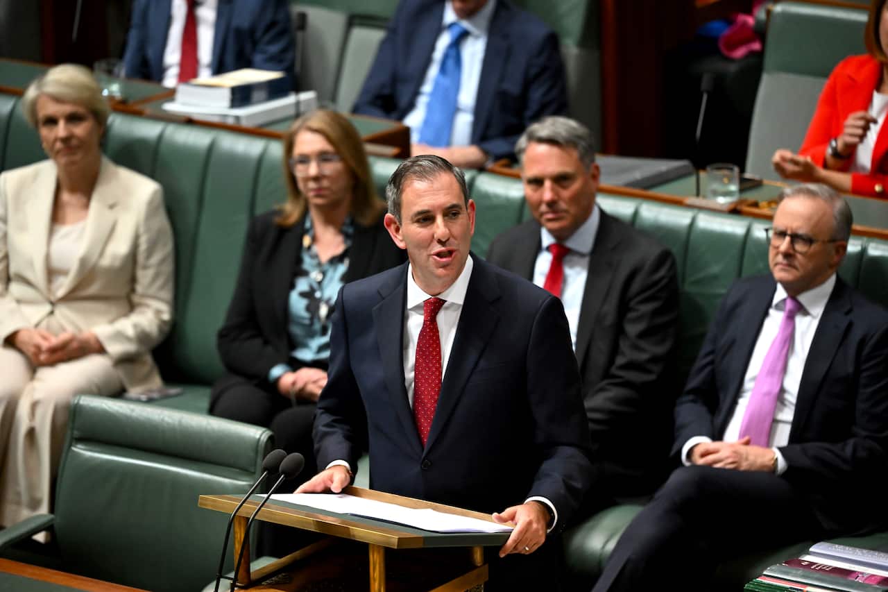 A man in a suit and tie speaks while other parliamentarians sit behind him