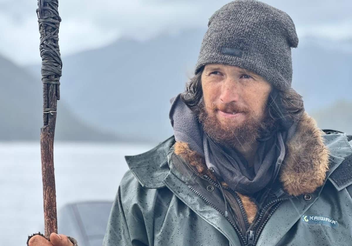 A man with matted facial hair, wearing a windbreaker, holding a stick.