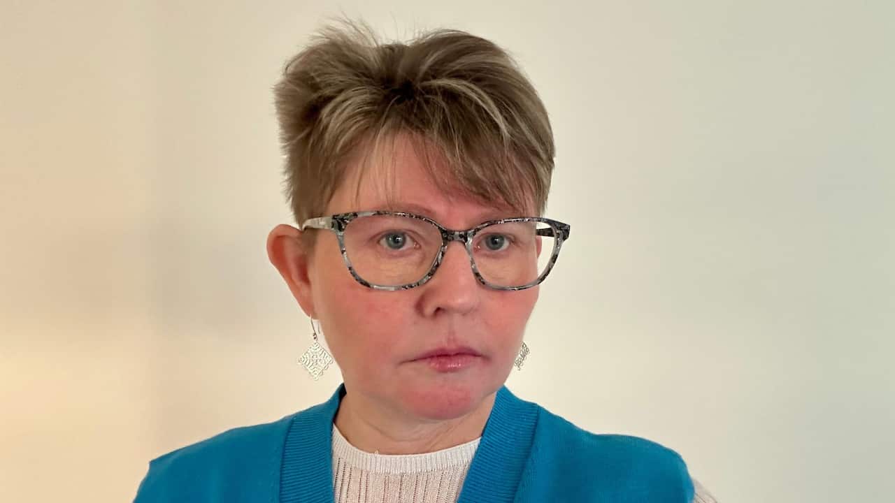 a middle aged woman with large glasses and short blonde hair in a blue jacket stands in front of a white wall