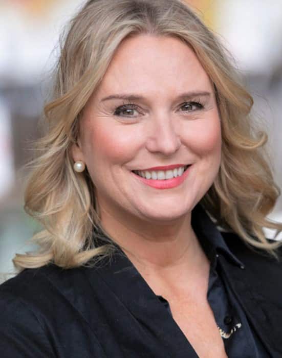 A woman with blonde hair and a black jacket smiles at the camera. 