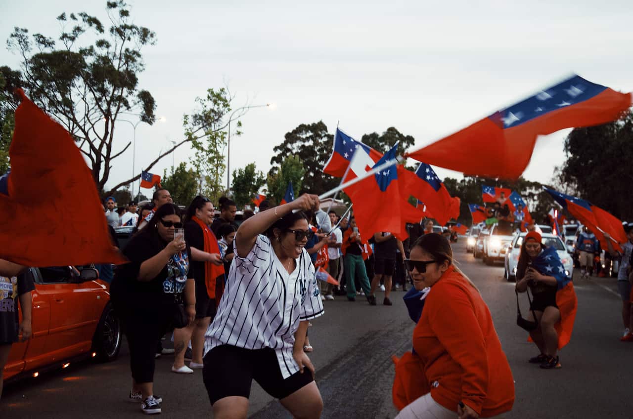 A group of women dance while waving the Samoan flag on the road. 