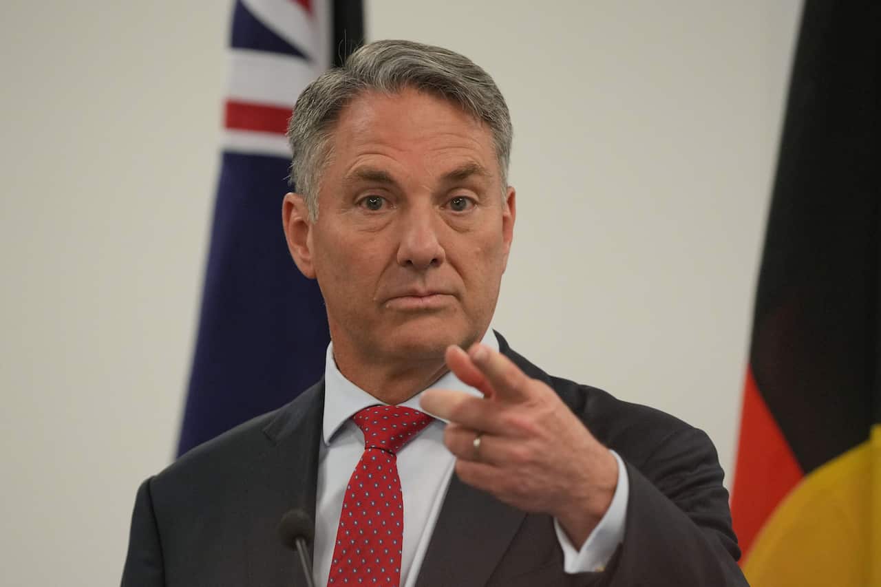 A man wearing a suit and a red tie points his finger as he speaks