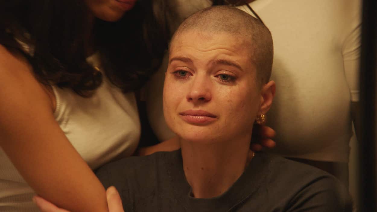 A young woman sits crying after having shaved her head. She is supported by two friends.jpg