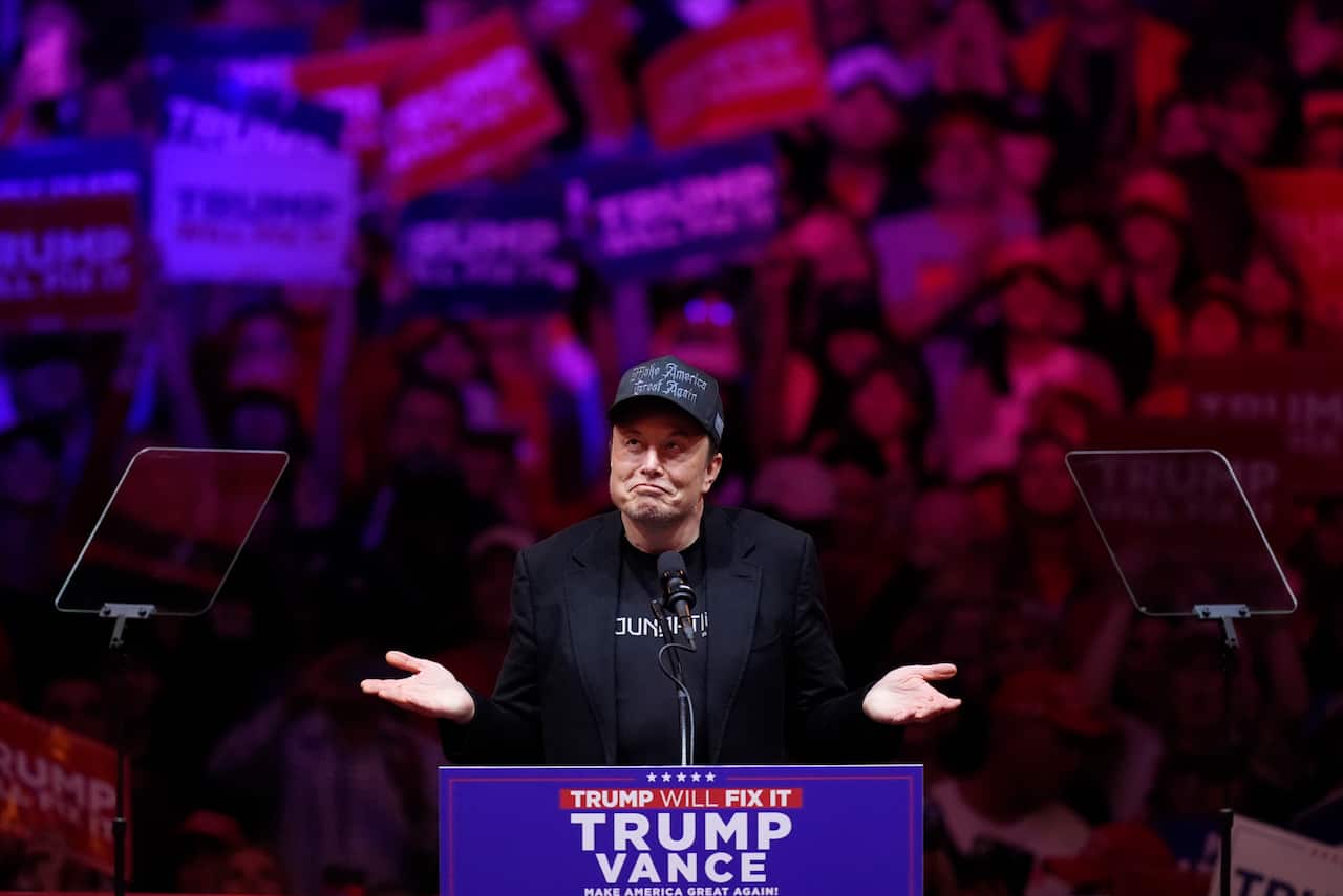 Elon Musk stands at a podium reading 'Trump Vance' wearing a dark suit and a baseball cap with an audience visible in the background