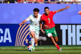 A footballer wearing a white shirt and shorts dribbles the ball while closely closely challenged by a defender.