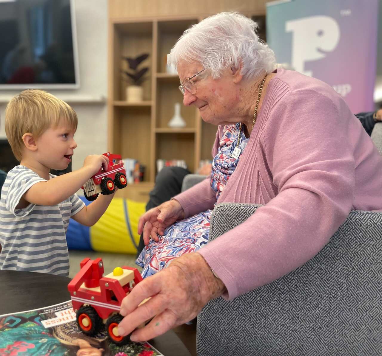 An older woman plays with a young boy holding a fire truck toy. 