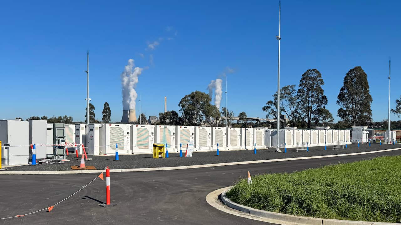 Rows of white cabinets containing batteries can be seen, while in the background two large cooling towers emit water vapour