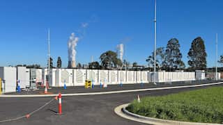 Rows of white cabinets containing batteries can be seen, while in the background two large cooling towers emit water vapour