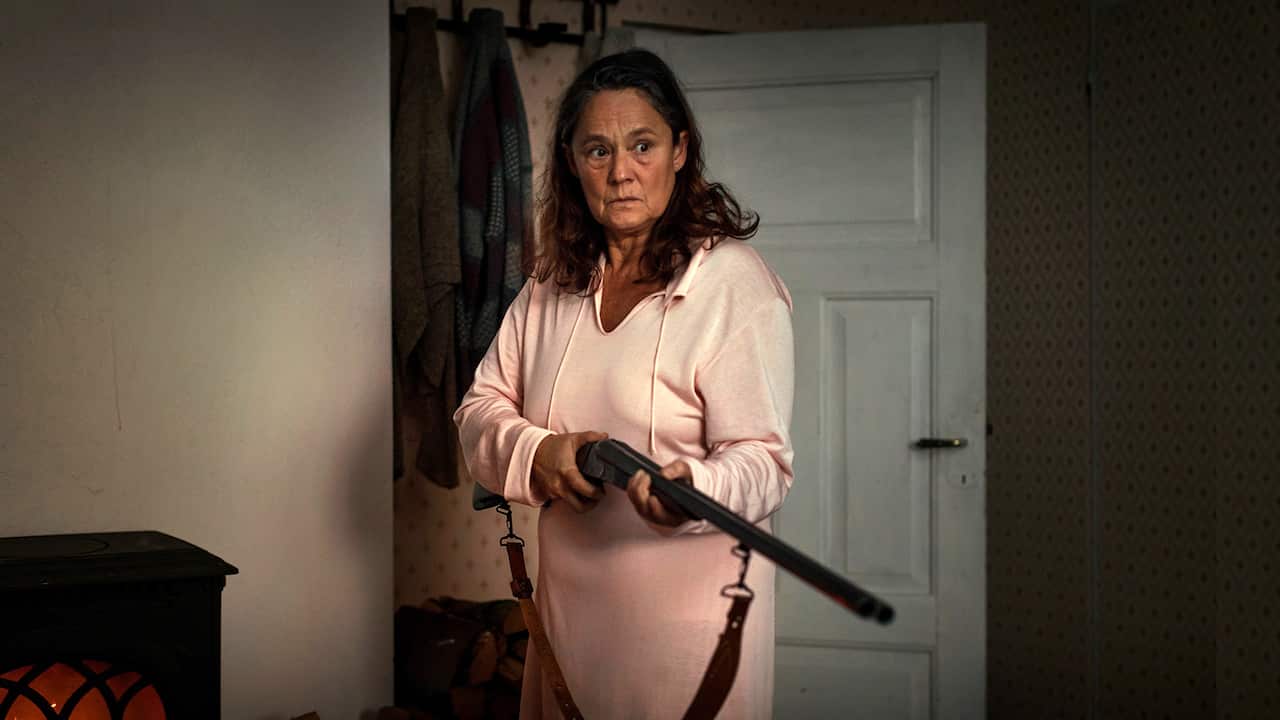 A worried woman stands in front of an open door, holding a riflge. 