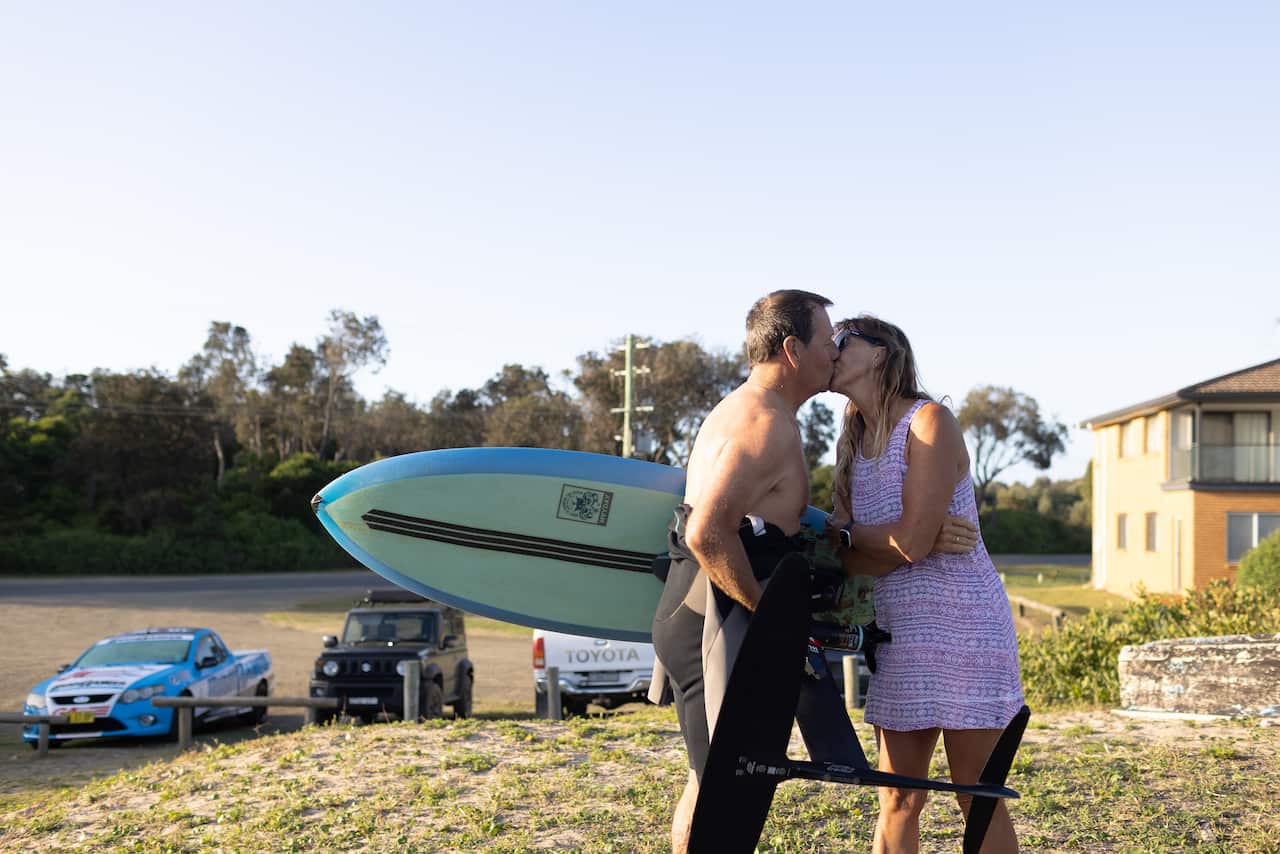 A couple kisses outdoors, with the man wearing a wetsuit and holding a surfboard, in front of a parking area with several vehicles and a house.