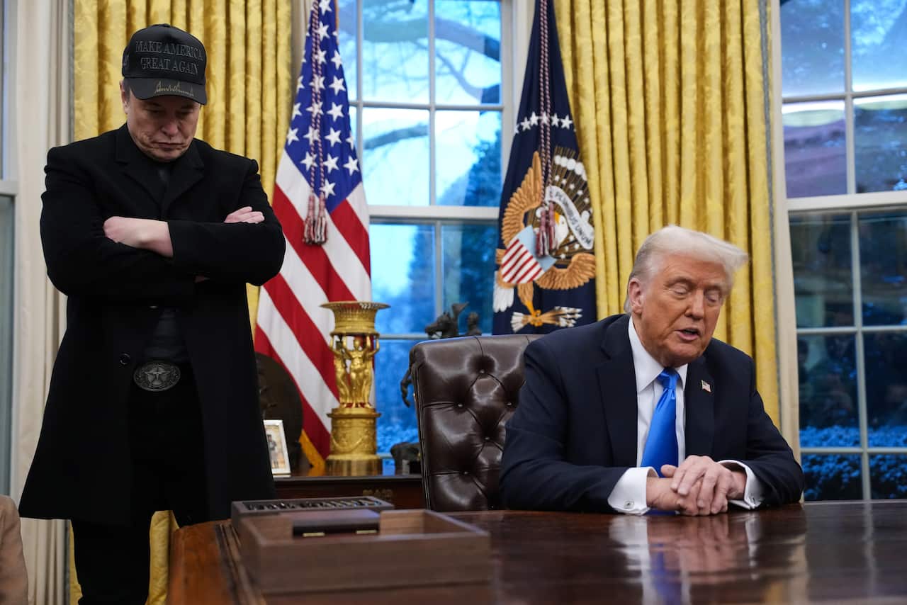 Elon Musk and Donald Trump in the Oval Office. Trump is sitting down, while Musk is standing near him, arms folded.