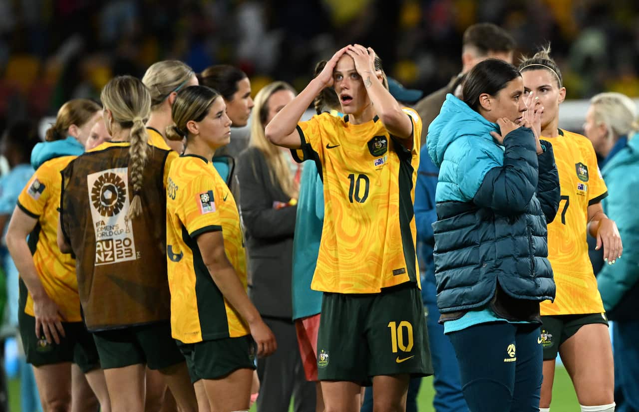 Group of Matildas players huddle together. Woman in middle of group holds her hands to the top of her face, with a shocked expression.