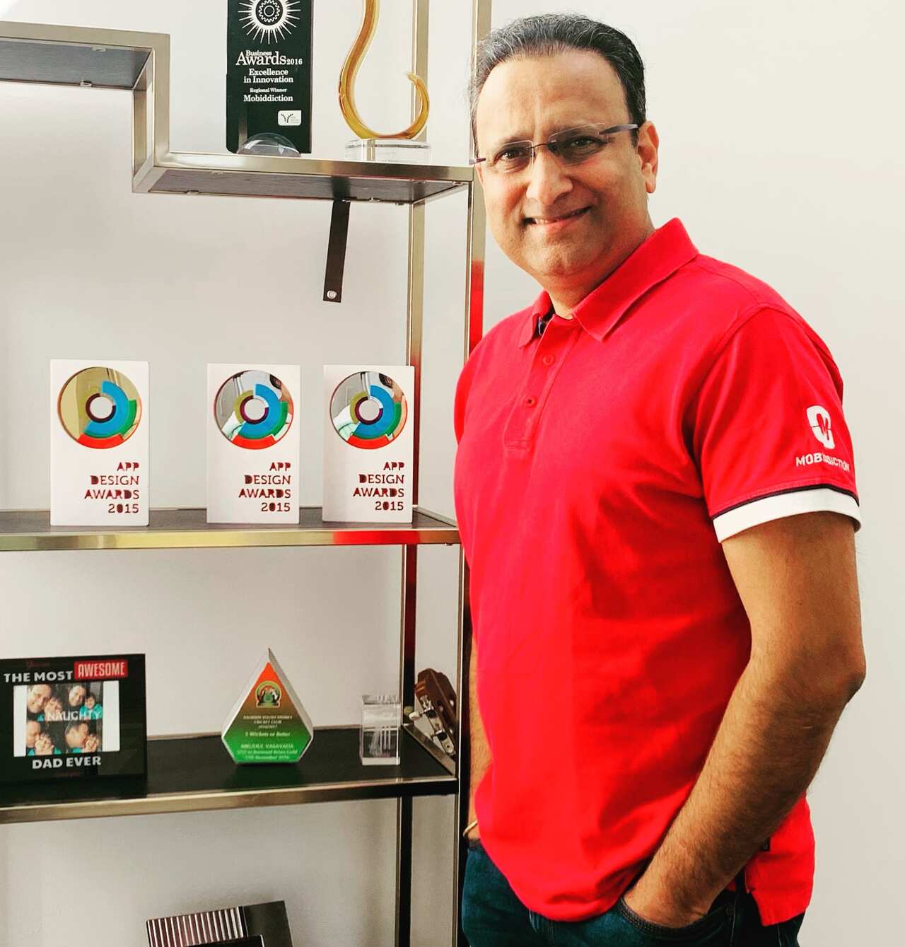 A man in a red t-shirt stands next to a shelf of awards.