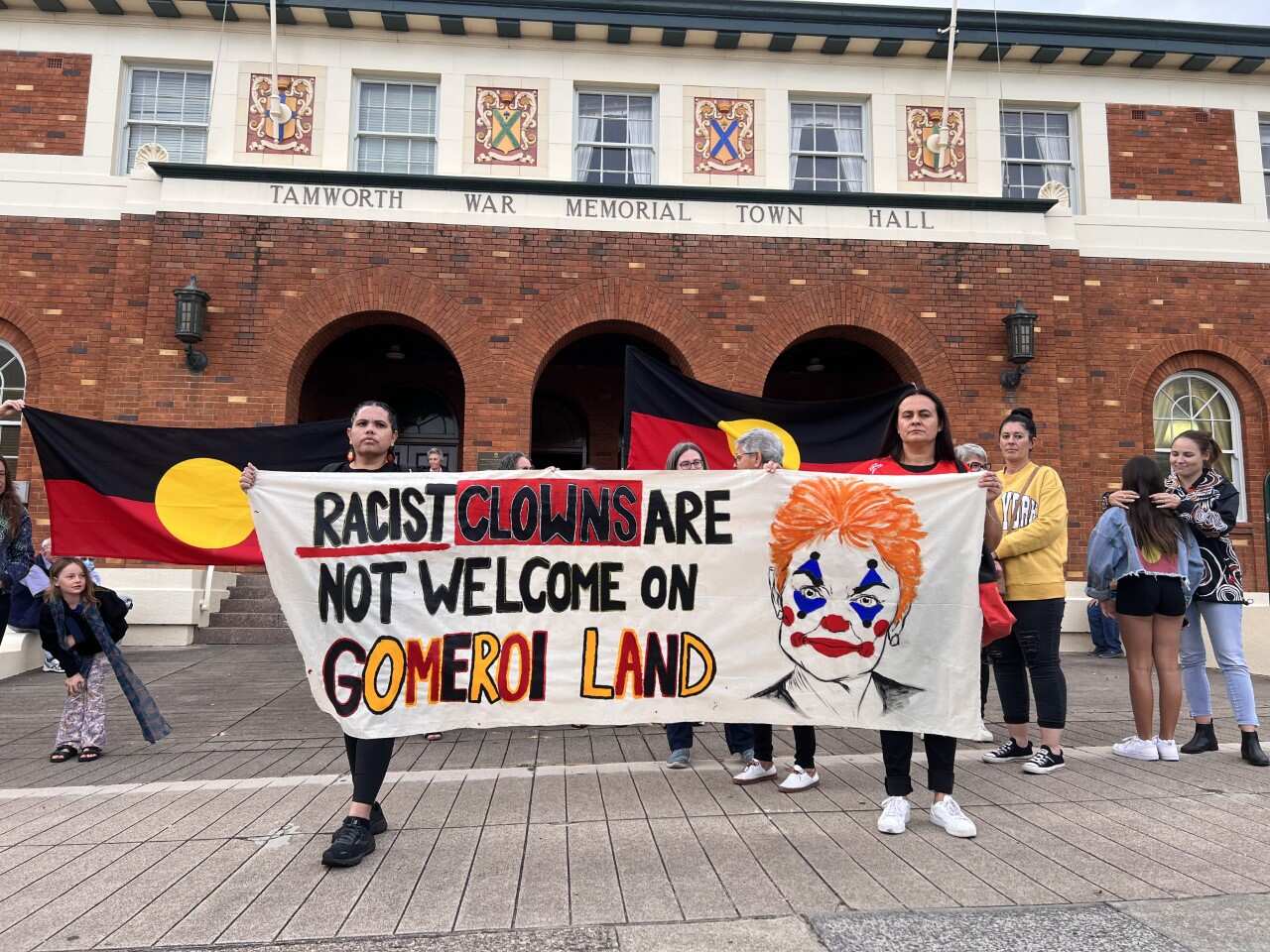 Protestors holding a banner with a red headed clown which reads: Racist clowns are not welcome on Gomeroi land.