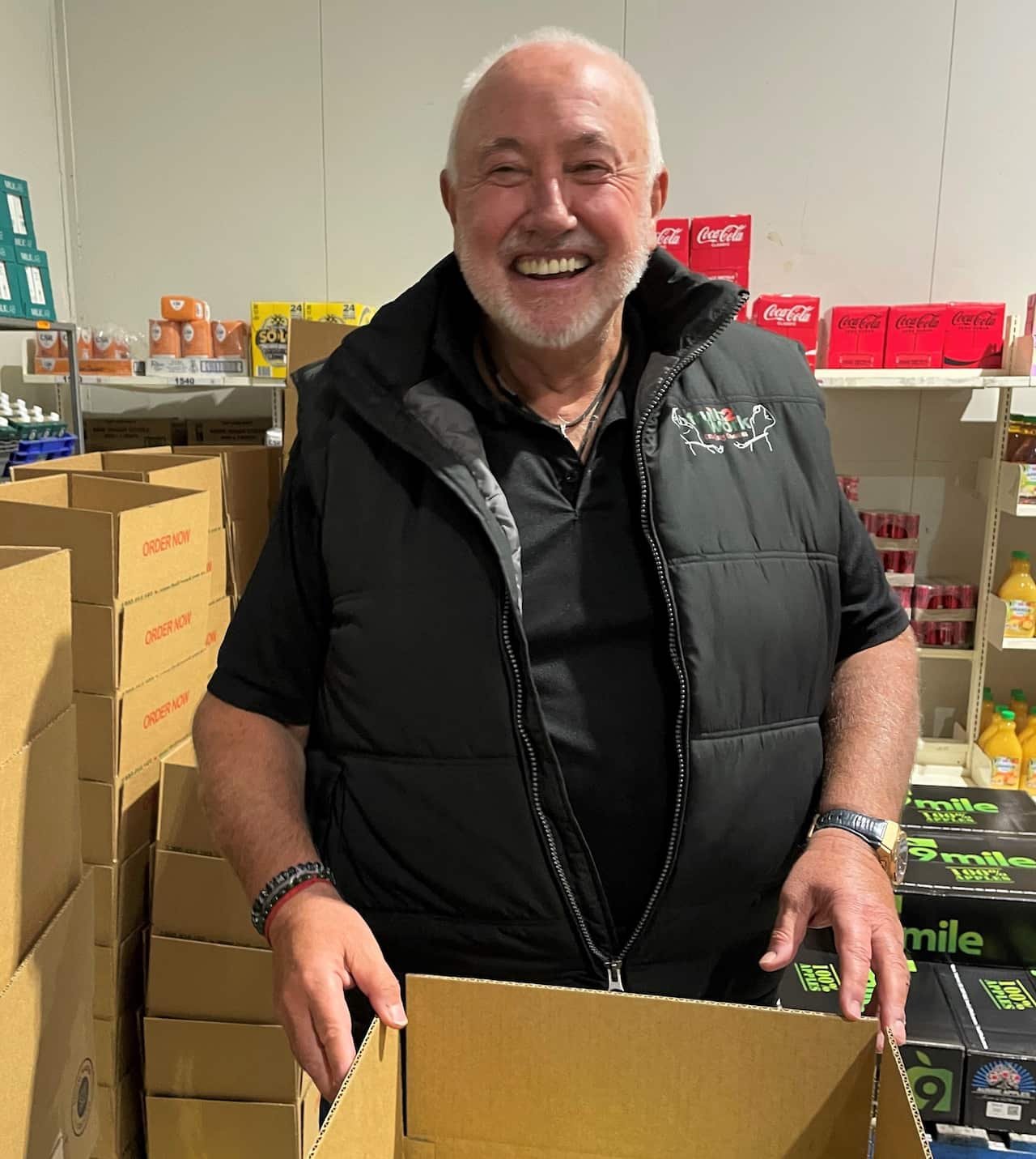 A man in a black vest stands in font of a cardboard box in a warehouse. 