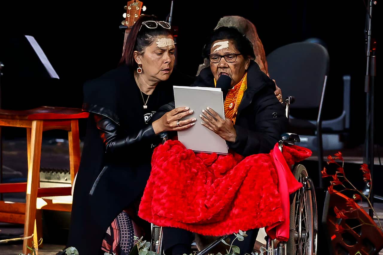 an old woman in a wheelchair reads into a microphone from prepared remarks, as another woman supports her. they both have traditional ochre smeared across their foreheads. 