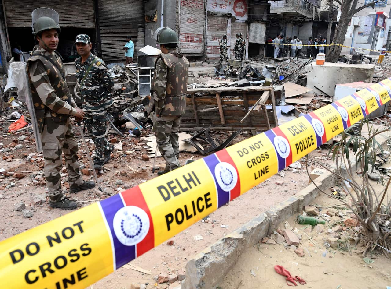 Yellow tape with red blue and white log markings reads, 'DO NOT CROSS LINE,' and 'DELHI POLICE'. Behind the tape are men in camoflage uniforms with helmets and shields. They stand on rubble of what appears to be destroyed and burned out buildings.